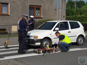 Technik kryminalistyki i dwie policjantki ruchu drogowego prowadzą oględziny miejsca wypadku drogowego. Obok przejścia dla pieszych stoi biały samochód i leży dziecięcy rowerek.