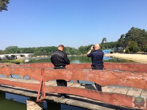 Funkcjonariusze stoją na moście nad  wodą, Policjant patrzy przez lornetkę, strażak notuje, w tle rozlewisko oraz plaża.