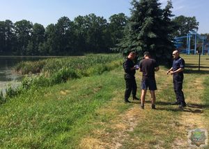 Policjant i strażak rozmawiają z mężczyzną stoją na trawiastym brzegu, od lewej strony fragment rozlewiska w tle drzewa, od prawej stalowa konstrukcja – zjeżdżalnia.