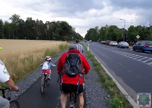 grupka rowerzystów zamykająca rajd jedzie po drodze rowerowej wzdłuż ul. Gorzowskiej, w oddali pierwsza część peletonu przejeżdża przez skrzyżowanie, policjanci zatrzymują ruch.