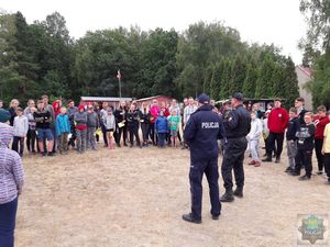 uczestnicy obozu harcerskiego stoją gromadą na wyschniętym trawniku  w półkolu  wewnątrz policjant i strażak, którzy mówią o bezpieczeństwie podczas wakacji. W tle niskie zabudowania, maszt z biało-czerwoną flagą, dalej ściana lasu.