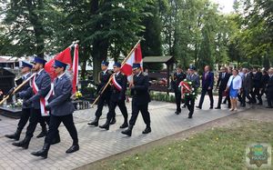 Poczet sztandarowy policji i straży pożarnej maszeruje cmentarną aleją, w tle zgromadzeni na uroczystości mieszkańcy.