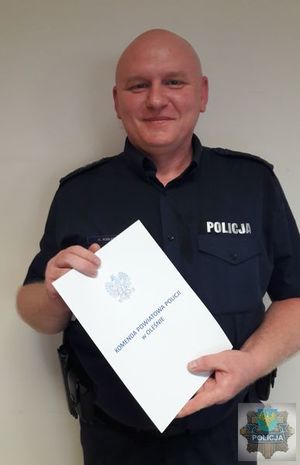 Policjant dzielnicowy  asp. sztab. Artur Kuligstoi  w umundurowaniu służbowym, trzyma w ręku list gratulacyjny, jest uśmiechnięty.