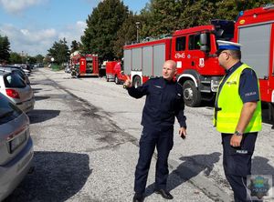 Parking przy szpitalu Rejonowym w Oleśnie - Komendant Powiatowy Policji w Oleśnie - inspektor Tomasz Kubicki przekazuje informację wskazując rejon szpitala Naczelnikowi Wydziału Prewencji i Ruchu Drogowego, w tle zaparkowane wozy bojowe Straży Pożarnej.