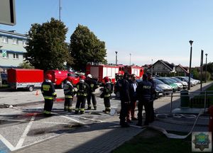 Parking przed głównym wejściem do szpitala, 6 strażaków w umundurowaniu bojowym rozmawiają między sobą, obok stoi 4 policjantów. Na chodniku i asfalcie rozwinięte węże gaśnicze. W tle po lewej stronie zaparkowane  Strażackie Wozy Bojowe, po prawej stronie samochody cywilne.