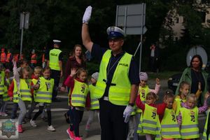 Policjant ruchu drogowego podnosi rękę i zatrzymuję ruch. Przedszkolaki przechodzą po pasach z podniesioną do góry ręką.