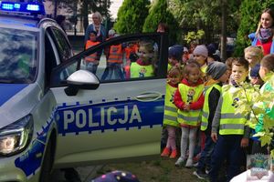 Przedszkolaki stoją obok radiowozu z otwartymi drzwiami. Siedzący w środku policjant prezentuje sygnały błyskowe i dźwiękowe.