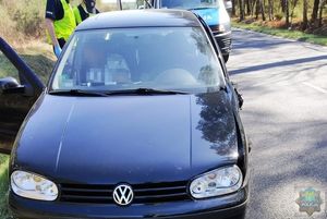 Policjanci w kamizelkach odblaskowych stoją za samochodem, który uczestniczył w kolizji drogowej. W tle stoi oznakowany radiowóz-bus.