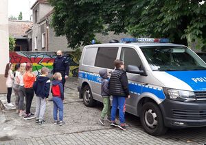 Dzieci stoją na zewnątrz budynku, patrzą na policjanta stojącego obok radiowozu. Dwóch chłopców podchodzi do pojazdu i zagląda przez szybę.