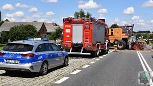 Policyjny radiowóz na miejscu wypadku drogowego. W tle widoczni policjanci i strażacy pracujący na miejscu zdarzenia oraz kombajn zbożowy