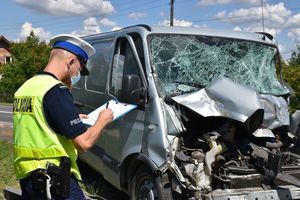 Policjant ruchu drogowego wypełnia protokół oględzin stojąc przed rozbitym samochodem dostawczym