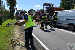 Funkcjonariusz ruchu drogowego wykonuje pomiary na miejscu wypadku drogowego. W tle kombajn i rozbity samochód dostawczy