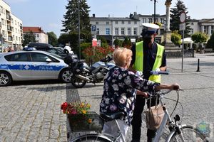 Policjant ruchu drogowego w kamizelce odblaskowej podaje rowerzystce odblask. W  tle widoczny radiowóz i policyjny motocykl