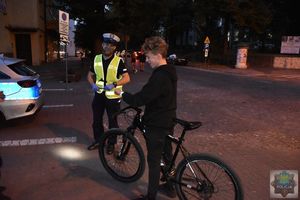 Policjant ruchu drogowego stoi obok rowerzysty, który prezentuje założoną na lewej ręce odblaskową opaskę.