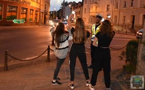 Policjant w żółtej odblaskowej kamizelce stoi obok trzech kobiet, które prezentują założone na ręce odblaskowe opaski.