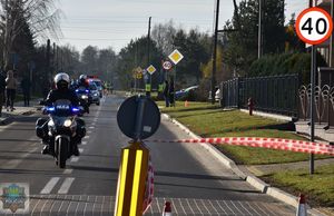 Policjanci na motorach jadą na czele wyścigu, za nimi biały pojazd transmisyjny za którym zza zakrętu wybiega czołówka wyścigu. Po lewej stronie na chodniku osoby kibicujące zawodnikom.