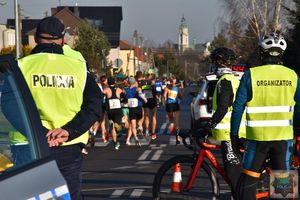 Policjant stoi na skrzyżowaniu,  za nim  peleton biegaczy, po prawej stronie osoby w kamizelkach z napisem organizator. 
W tle na dalekim planie wieża kościoła.