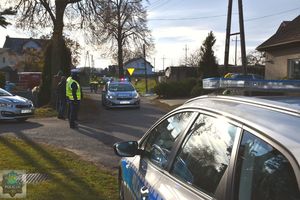 Stojące radiowozy na pierwszym  planie oraz po stronie lewej, stojący policjant. Dalej dojeżdżający radiowóz pilotujący zawodników.