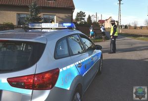 Radiowóz policyjny ustawiony przed skrzyżowaniem za pojazdem stoi policjant , obok przebiega dwóch zawodników.