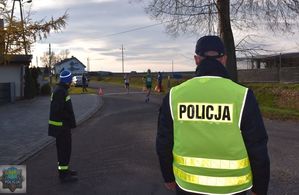 Policjant stoi na skrzyżowaniu , obok Strażak z Ochotniczej Straży Pożarnej, za nimi  dobiegający zawodnicy, w tle zabudowania , pola i niebo.