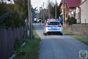 wiejski odcinek drogi asfaltowej widoczne zabudowania, radiowóz -   oddala się jadąc  za biegaczami.