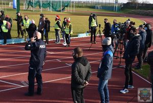 Fragment bieżni lekkoatletycznej - dziennikarze oraz służby organizatora zawodów stoją za metą maratonu. Komendant Powiatowy Policji - insp. Tomasz Kubicki obserwuje finiszujących zawodników trzyma przy uchu telefon.