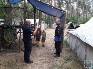 Policjant  w towarzystwie Strażaka stoją przed prowizoryczną drewnianą  bramą do leśnego obozowiska harcerskiego. Dwoje dzieci w mundurkach harcerskich wita uniesionymi dłońmi funkcjonariuszy. Z prawej strony widoczny namiot biwakowy. W tle drzewa.