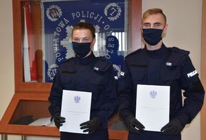 Nowomianowani policjantka i policjant pozują do zdjęcia. W rękach trzymają akty mianowania na  stopień posterunkowego policji. W tle sztandar jednostki.