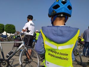 Dziecko w kasku i kamizelce odblaskowej z napisem z myślą o twoim bezpieczeństwie. Na drugim planie policjant w kasku i białej koszulce z napisem policja.